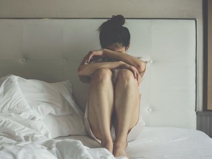 Junge Frau in hellem Nachthemd sitzt verzweifelt mit angezogenen Knien auf einem ungemachten Bett.