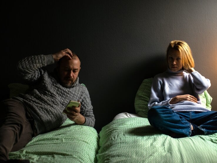 Gelangweiltes oder distanziertes Paar, das in getrennten Betten sitzt. Der Mann schaut auf sein Handy, während er sich am Kopf kratzt, und die Frau sitzt nachdenklich auf ihrem Bett. | ©  Adobe Stock / Konstiantyn Zapylaie