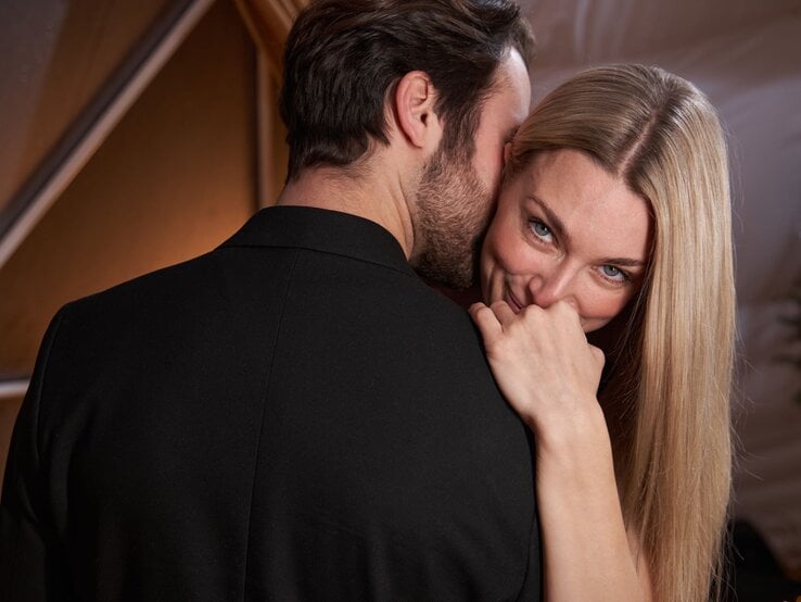 Eine blonde Frau mit langem Haar lächelt verschmitzt, während ein Mann im dunklen Anzug ihr leise etwas ins Ohr flüstert.