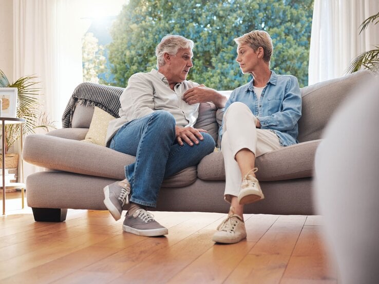 Ein älteres Paar sitzt auf einem beigen Sofa im sonnendurchfluteten Wohnzimmer und führt ein ernstes Gespräch.