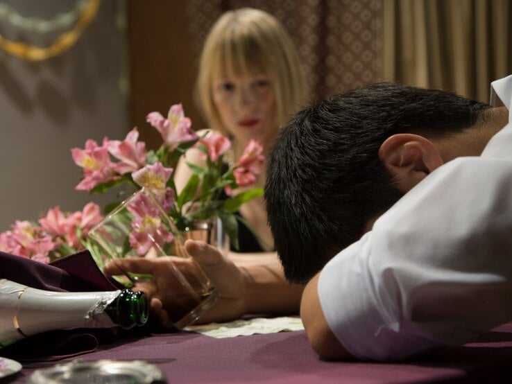 Ein Mann in weißem Hemd liegt mit dem Kopf auf einem Tisch, ein leeres Glas in der Hand, während eine blonde Frau ihn ernst ansieht.