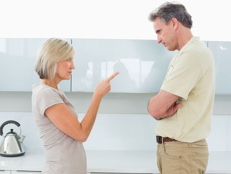 Eine blonde Frau in beigem T-Shirt zeigt vorwurfsvoll mit dem Finger auf einen Mann in heller Kleidung, der mit verschränkten Armen abwehrend blickt.