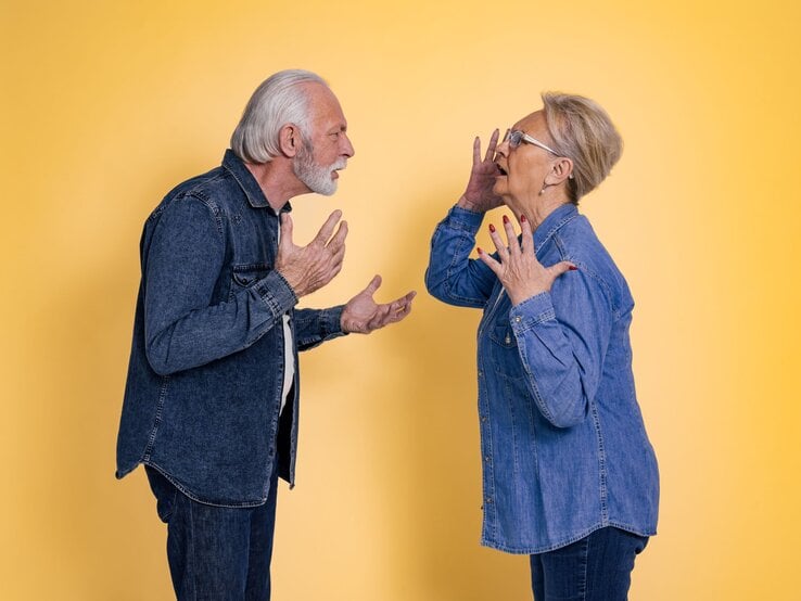 Älteres Paar in Jeanskleidung streitet heftig vor gelbem Hintergrund, beide gestikulieren mit aufgebrachten Gesichtern.