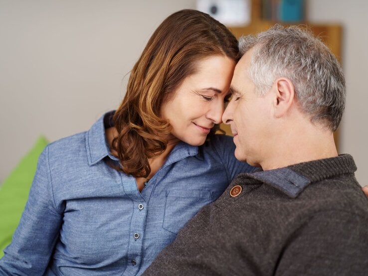 Eine braunhaarige Frau und ein grauhaariger Mann sitzen innig umarmt und lächelnd, Stirn an Stirn, auf einem Sofa.