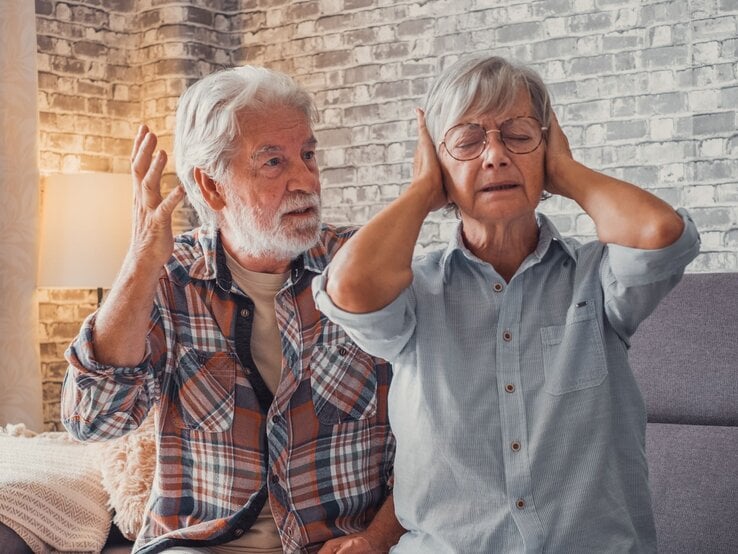 Älterer Mann gestikuliert verärgert, während eine ältere Frau mit geschlossenen Augen die Ohren zuhält und genervt wirkt.