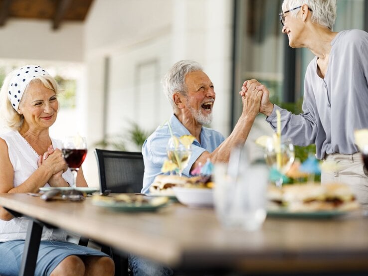 Drei ältere Menschen lachen und feiern an einem gedeckten Tisch im Freien, während zwei sich herzlich die Hände reichen.