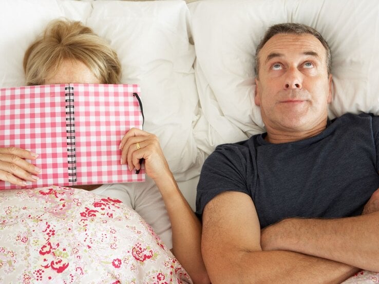 Mittelaltes Paar liegt im Bett, die Frau verbirgt ihr Gesicht hinter einem karierten Notizbuch, der Mann blickt genervt zur Decke.