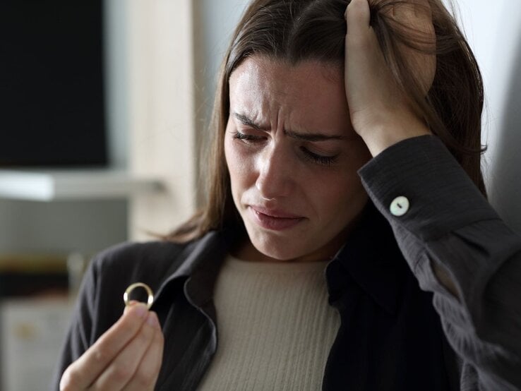 Junge Frau mit langen braunen Haaren blickt traurig auf einen Ring in der Hand, stützt den Kopf verzweifelt mit der anderen.