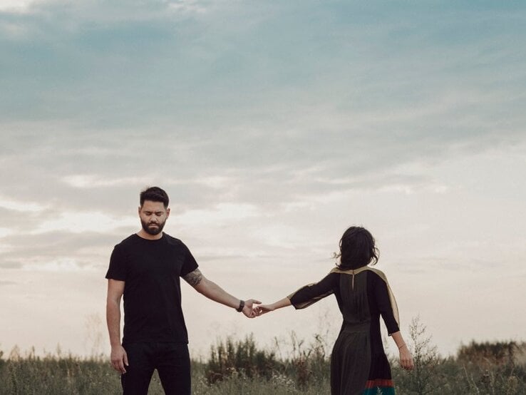 Ein bärtiger Mann in Schwarz hält traurig die Hand einer Frau im Kleid, die sich abwendet, vor wolkigem Himmel auf dem Feld.