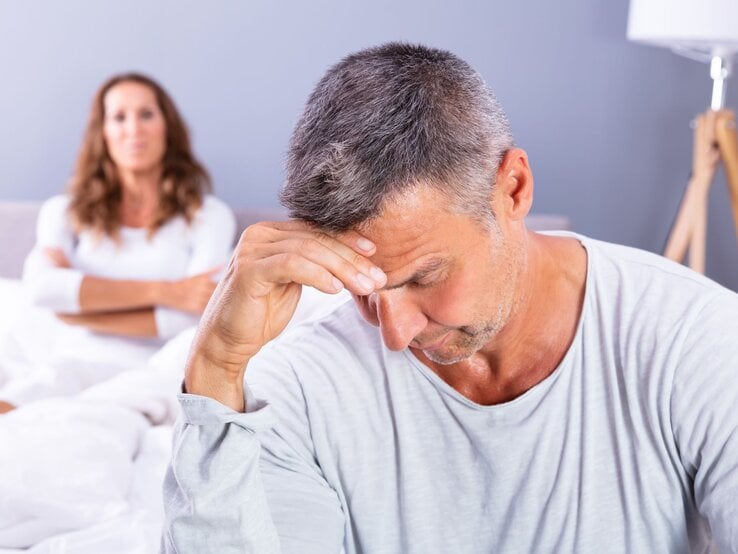 Ein nachdenklicher Mann mit grauem Haar sitzt bedrückt am Bettrand, im Hintergrund blickt eine Frau mit verschränkten Armen ernst.