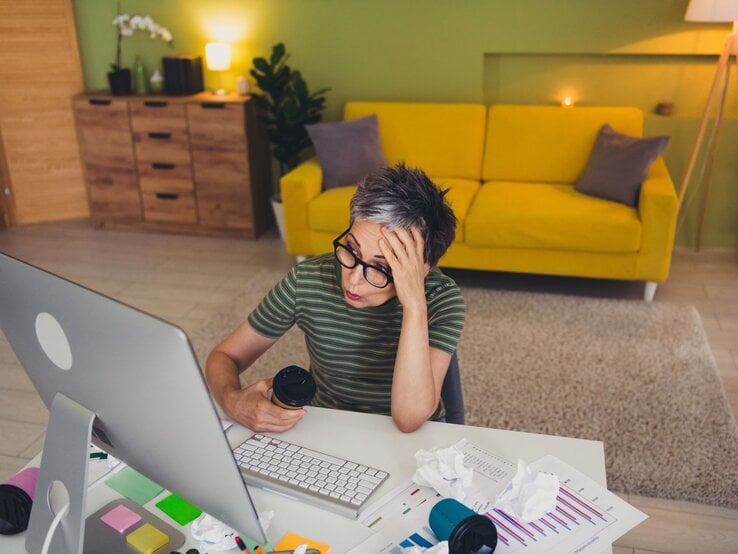 Eine gestresste Frau mit kurzer grauer Frisur sitzt mit Kaffeebecher vor Papieren am Schreibtisch in einem bunten Wohnzimmer.