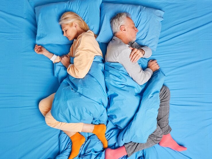 Eine blonde Frau und ein grauhaariger Mann schlafen Rücken an Rücken in farbigen Pyjamas auf blau bezogenem Bett.