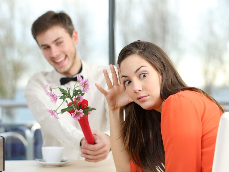 Junge Frau mit langen braunen Haaren blickt genervt zur Seite, während ein junger Mann ihr lachend einen Blumenstrauß hinhält. | © Getty Images / AntonioGuillem