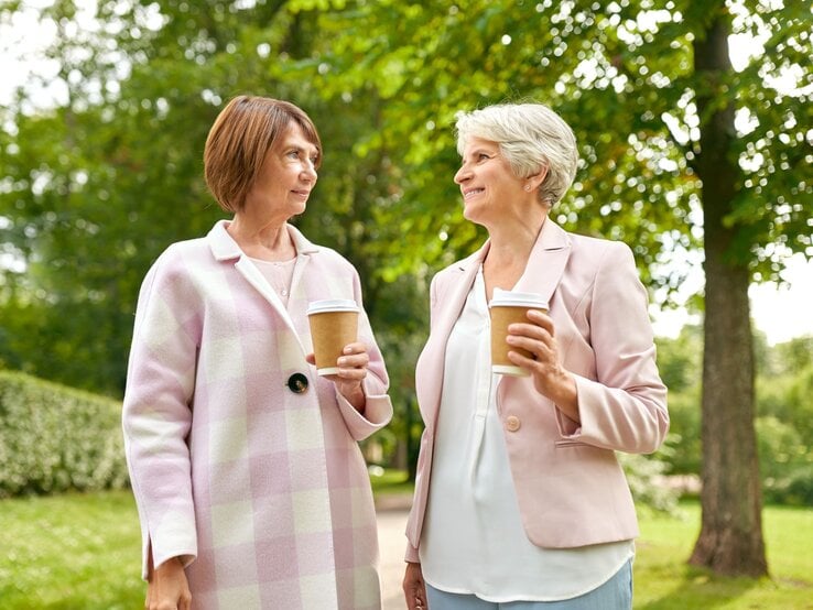 Zwei reife Frauen mit kurzen Frisuren unterhalten sich entspannt mit Coffee-to-go-Bechern in einem sonnigen Park mit Bäumen.