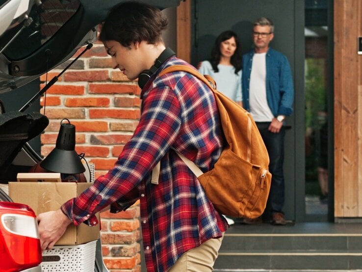Junger Mann mit Kurzhaarschnitt packt Kartons in Kofferraum, trägt kariertes Hemd und Rucksack, Eltern blicken gerührt aus Haustür.