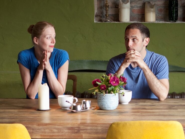Junge Frau und junger Mann in blauen Shirts sitzen sich mit verschränkten Händen an einem Holztisch gegenüber und schauen sich skeptisch an.