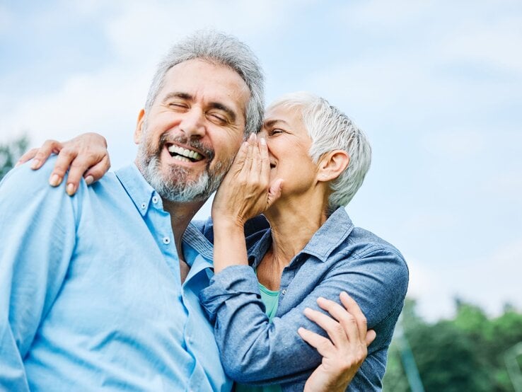 Grauhaariger, lachender Mann in hellblauem Hemd, dem eine kurzhaarige Frau liebevoll ins Ohr flüstert, vor blauem Himmel.