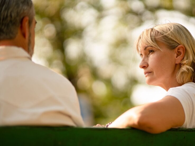 Blonde, nachdenkliche Frau mittleren Alters hört auf einer Parkbank einem Mann mit grauem Haar bei warmem Sonnenlicht zu.
