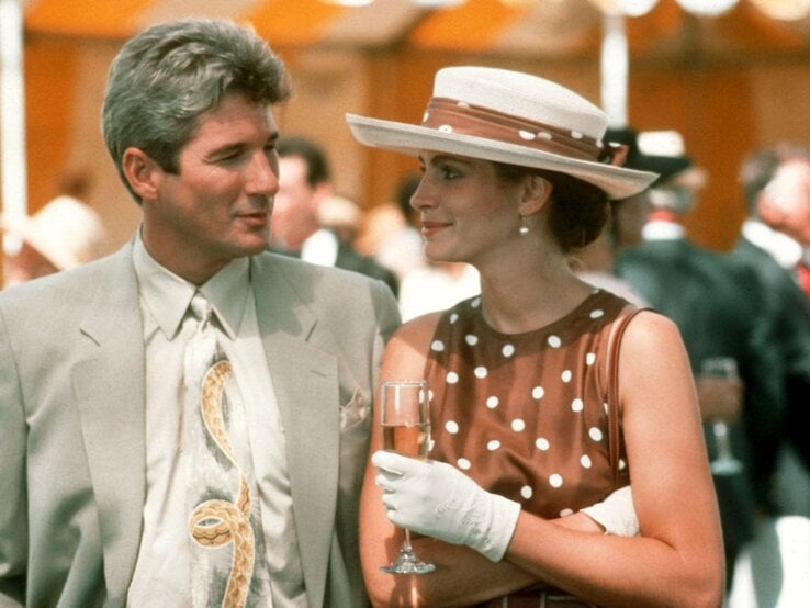 Eleganter Mann im hellen Anzug blickt lächelnd auf junge Frau im braunen Punktekleid mit Sektglas und weißem Hut bei Empfang.