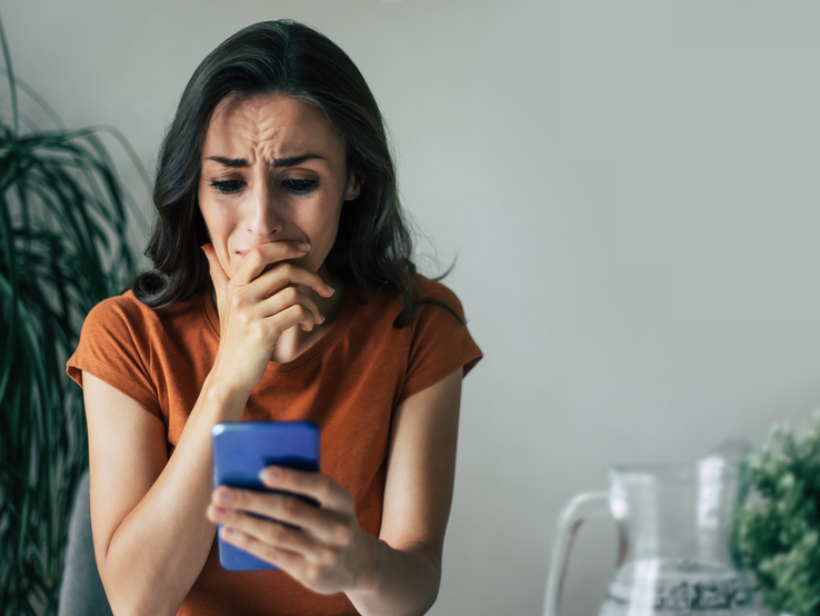Besorgte Frau in orangefarbenem Shirt liest fassungslos eine Nachricht auf dem Smartphone, im Hintergrund grüne Zimmerpflanze.