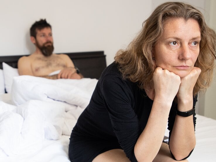 Eine Frau sitzt mit nachdenklicher Haltung im schwarzen Kleid auf einem weißen Bett, im Hintergrund ein Mann auf der Matratze.