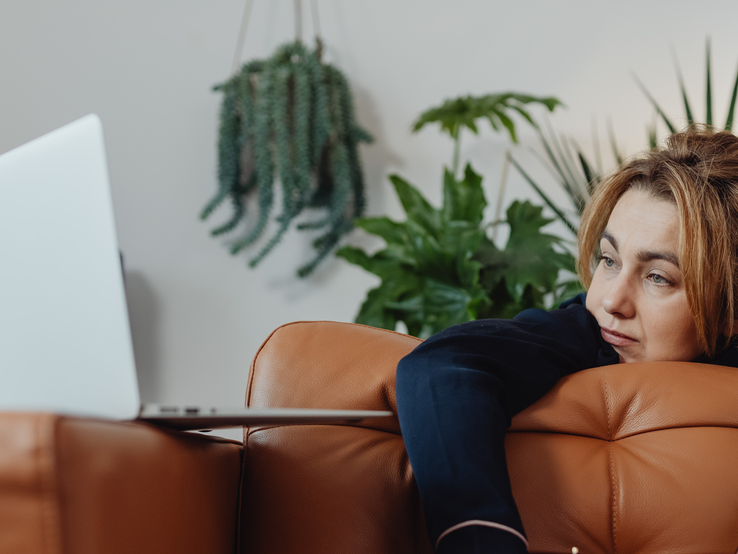 Eine Person lehnt mit dunkelblauem Ärmel entspannt auf einem braunen Ledersofa, im Hintergrund grüne Zimmerpflanzen und ein Laptop.