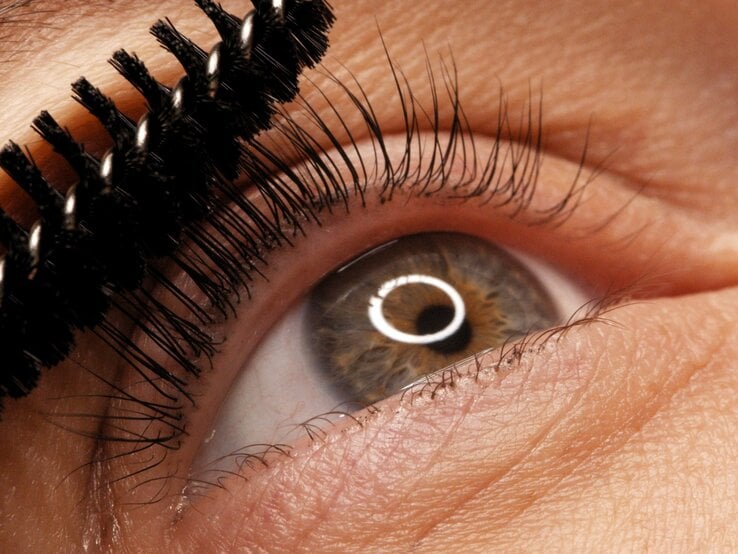 Nahaufnahme eines Auges mit brauner Iris, das mit einer Mascara-Bürste die oberen Wimpern tuscht.