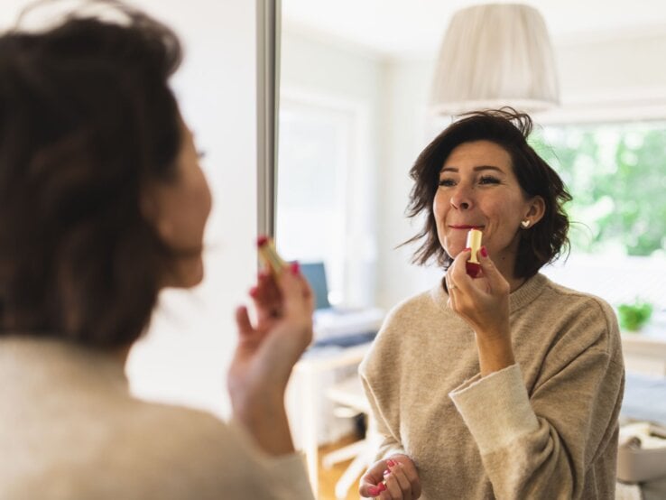 Frau im beigen Pullover trägt Lippenstift vor einem Spiegel auf, im Hintergrund ein gemütliches, gut beleuchtetes Wohnzimmer.