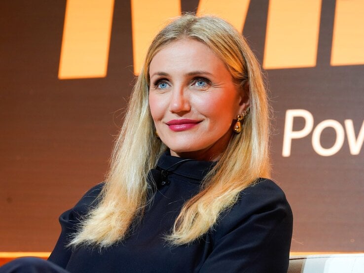 Cameron Diaz mit langen, offenen Haaren und goldenen Ohrringen lächelt dezent vor einem orange-schwarzen Hintergrund.