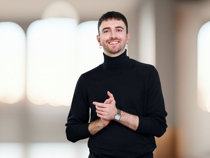 Junger Mann mit dunklem Kurzhaarschnitt, schwarzem Rollkragenpullover und Ohrring lächelt freundlich in modernem Innenraum. | © Frederic Schuckert