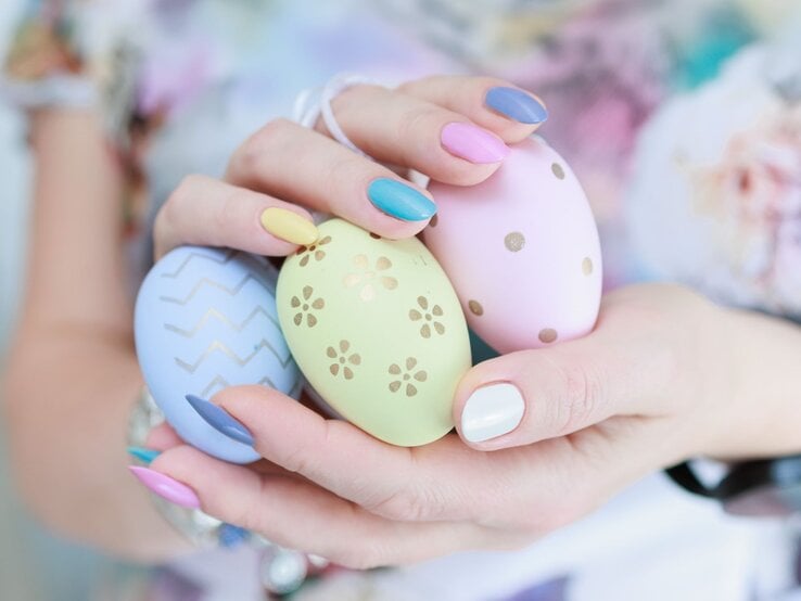 Hände einer jungen Frau mit pastellfarbenem Nageldesign halten drei bemalte Ostereier mit goldenen Mustern vor bunter Kleidung.