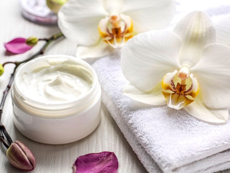 Eine luxuriöse Anti-Aging-Creme in Glasdose liegt auf einem weißen Handtuch, umgeben von Orchideen und Blütenblättern.