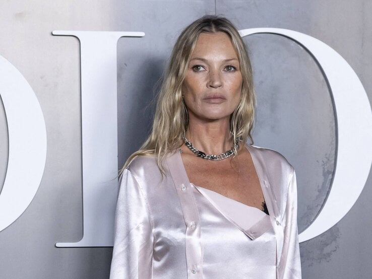 Kate Moss mit welligem Haar trägt ein seidiges, hellrosafarbenes Outfit und eine silberne Kette vor grauem Hintergrund.