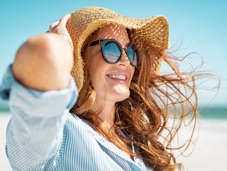 Rothaarige Frau in gestreifter Bluse genießt den Tag am Meer, eingecremt mit Sonnencreme und lächelt in die Sonne, während ihr Haar lebhaft flattert.