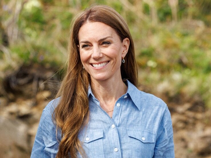 Lachende Prinzessin Kate mit langen braunen Haaren in hellblauer Bluse steht im Grünen vor verschwommenem Waldboden.