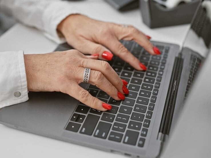 Gepflegte Frauenhände mit rotem Nagellack und silbernem Ring tippen auf einer Laptop-Tastatur in modernem Büroambiente.