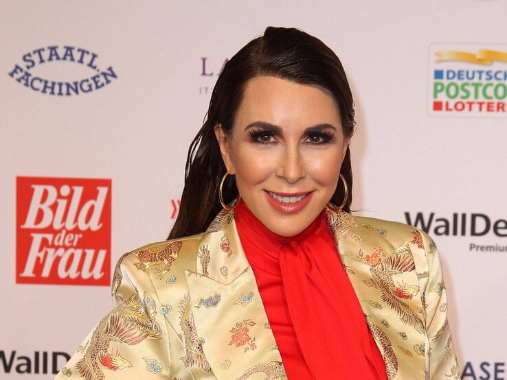 Judith Williams mit langen dunklen Haaren in goldener Seidenjacke mit Drachenmotiv und roter Bluse vor Fotowand mit Logos.