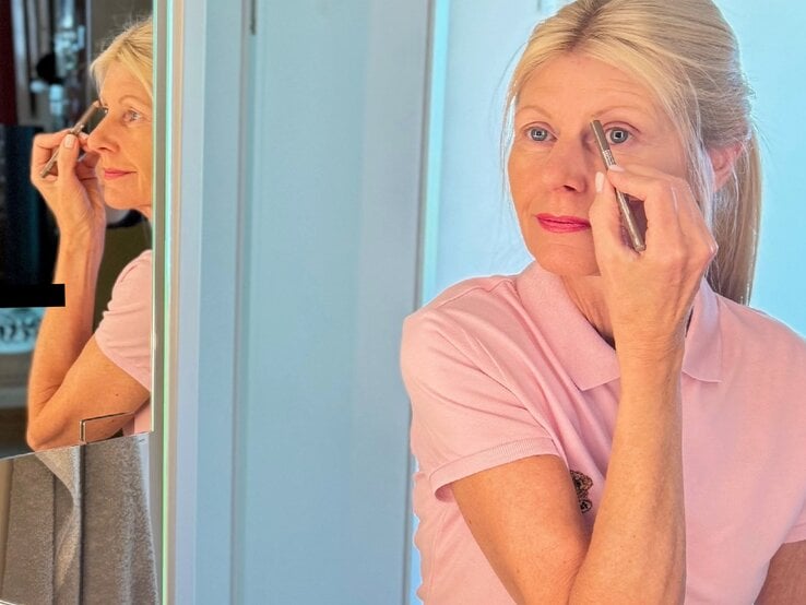 Blonde Renate Zott im mittleren Alters trägt konzentriert Augen-Make-up auf, ihr Spiegelbild ist links im hellen Badezimmer zu sehen.