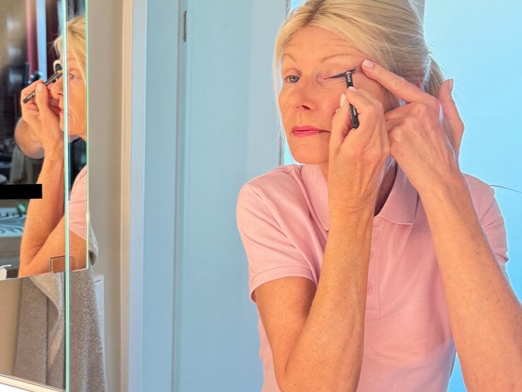 Blonde Renate Zott mittleren Alters im rosa Poloshirt schminkt sich am hellen Spiegel präzise mit schwarzem Kajalstift. | © Renate Zott