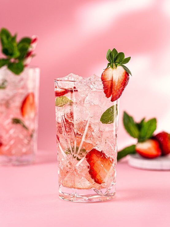 Kristallglas mit Erdbeer-Minz-Drink und Crushed Ice vor pinkem Hintergrund, garniert mit halber Erdbeere am Glasrand. | © Iryna Melnyk/ Glow25