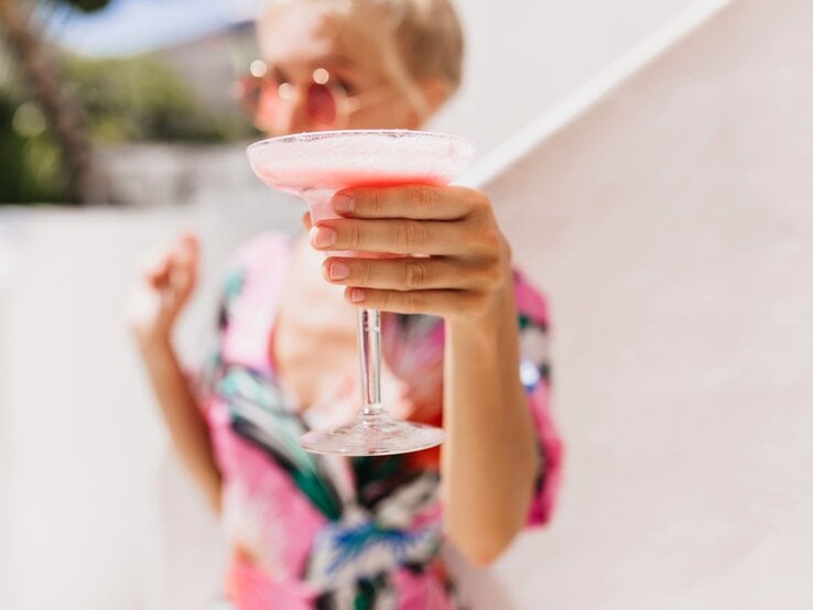 Verschwommenes Porträt einer Frau mit Sonnenbrille, die einen rosa Cocktail in die Kamera hält, vor heller Kulisse.