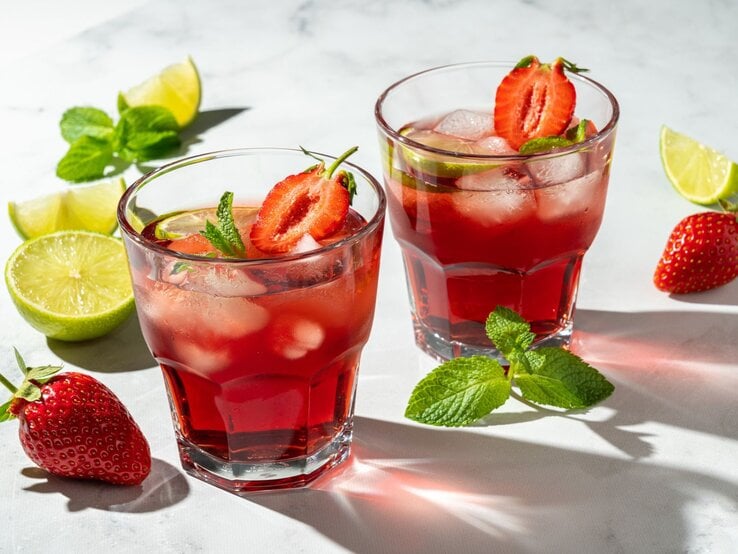 Eiskalte rote Cocktails mit halben Erdbeeren, Limettenstücken und Minzblättern leuchten im Sonnenlicht auf weißem Untergrund. | © Getty Images/ Natalia Gdovskaia