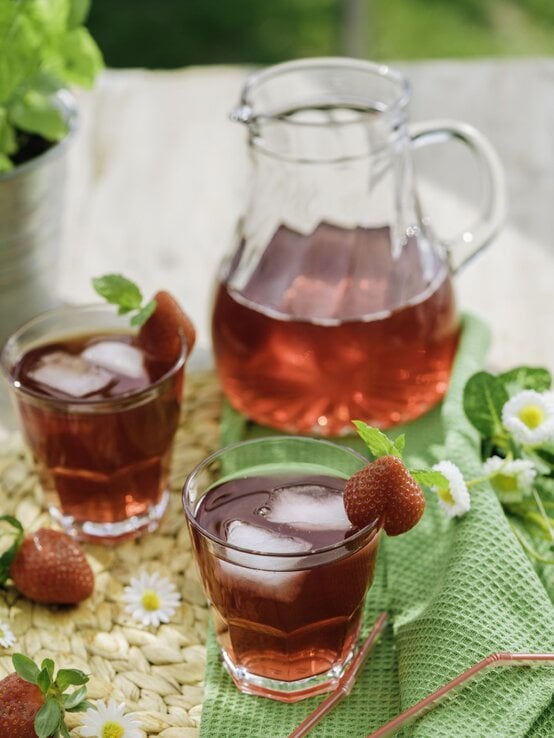Erdbeer-Eistee mit Eis und frischer Deko in Gläsern auf Bastmatte, daneben Gänseblümchen, Minze und eine Karaffe. | © Getty Images/ GMVozd