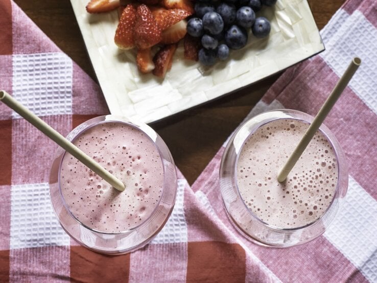 Zwei rosafarbene Smoothies mit Trinkhalm stehen auf rot-weißem Küchentuch, daneben ein Obstteller mit Beerenmix. | © Getty Images/ Monica Bertolazzi