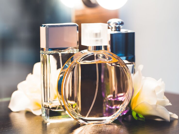 Drei elegante Parfümflakons in verschiedenen Formen stehen auf einem dunklen Tisch, umgeben von weißen Blumen und warmem Licht.