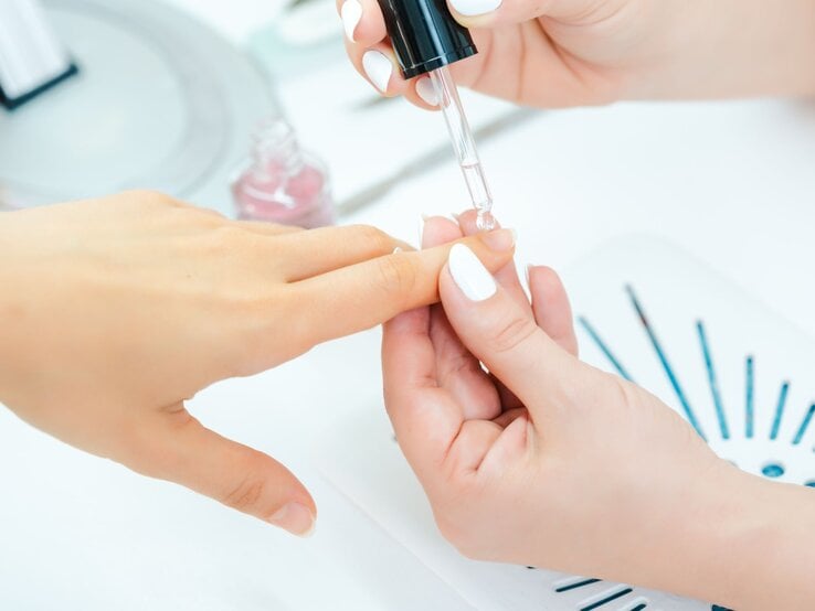 Nahaufnahme einer klassischen Maniküre: Nagelöl wird präzise mit einer Tropfpipette auf den Nagel einer Hand aufgetragen.