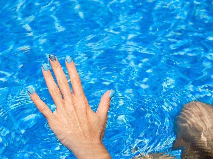 Hand mit blau lackierten Nägeln und glitzerndem Akzentfinger taucht in klares, funkelndes Poolwasser bei sonnigem Licht.
