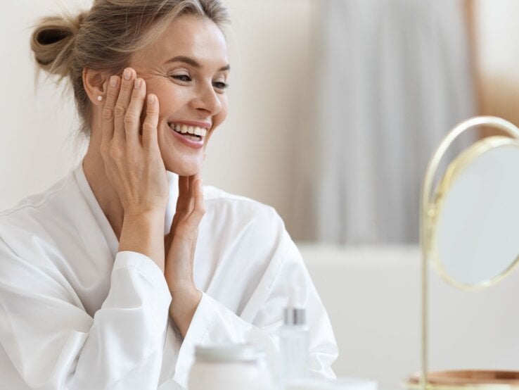 Frau in weißem Bademantel berührt sanft ihr Gesicht, vor einem Tisch mit Creme und einem goldenen Kosmetikspiegel im hellen Raum.