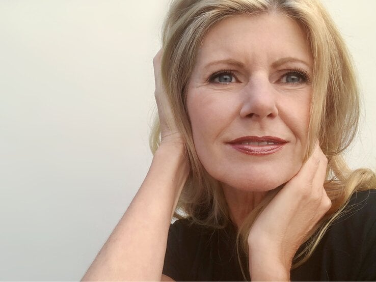 Eine Frau mit hellem, welligem Haar trägt ein schwarzes Oberteil und ruht mit einem Arm locker an ihrem Gesicht vor hellem Hintergrund.