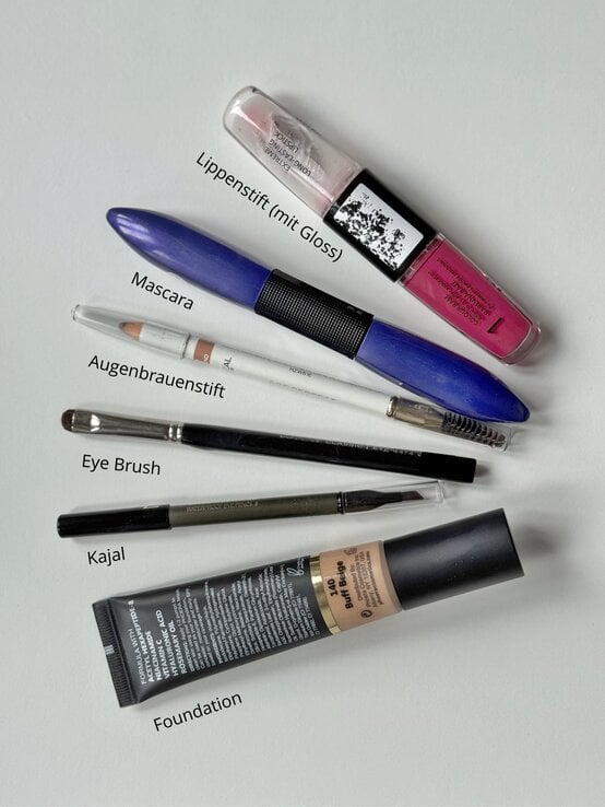 Verschiedene Make-up-Produkte, darunter Lippenstift, Mascara, Augenbrauenstift, Kajal, Pinsel und Foundation, liegen auf einer weißen Fläche | © Renate Zott